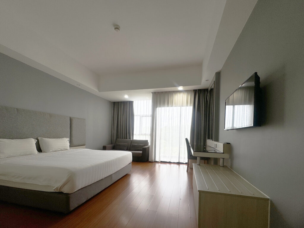 Interior of Deluxe King Room at Livingston Hotel Sandakan showing bed, TV, sofa, balcony, curtains, and luggage area