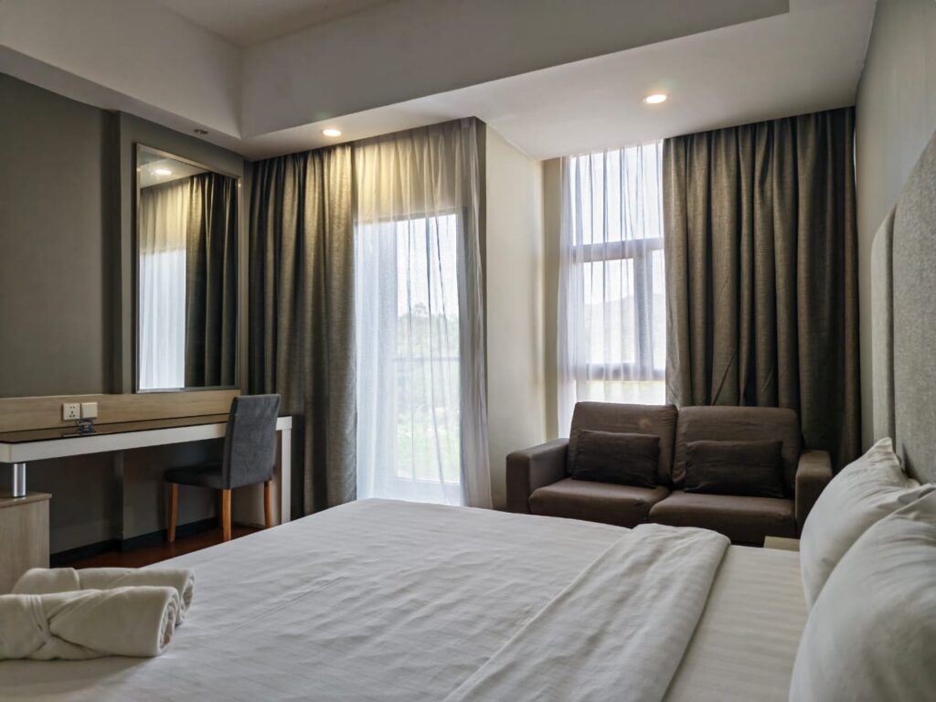Accessible room with bed, sofa, balcony, chair, work desk with mirror, and curtains at Livingston Hotel Sandakan Sabah