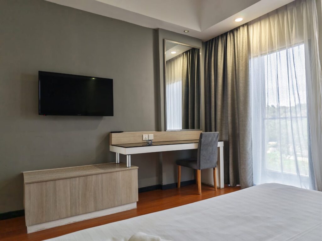 TV, mirror, desk with chair, luggage storage area, curtain, and balcony in Accessible (OKU) Room at Livingston Hotel Sandakan Sabah